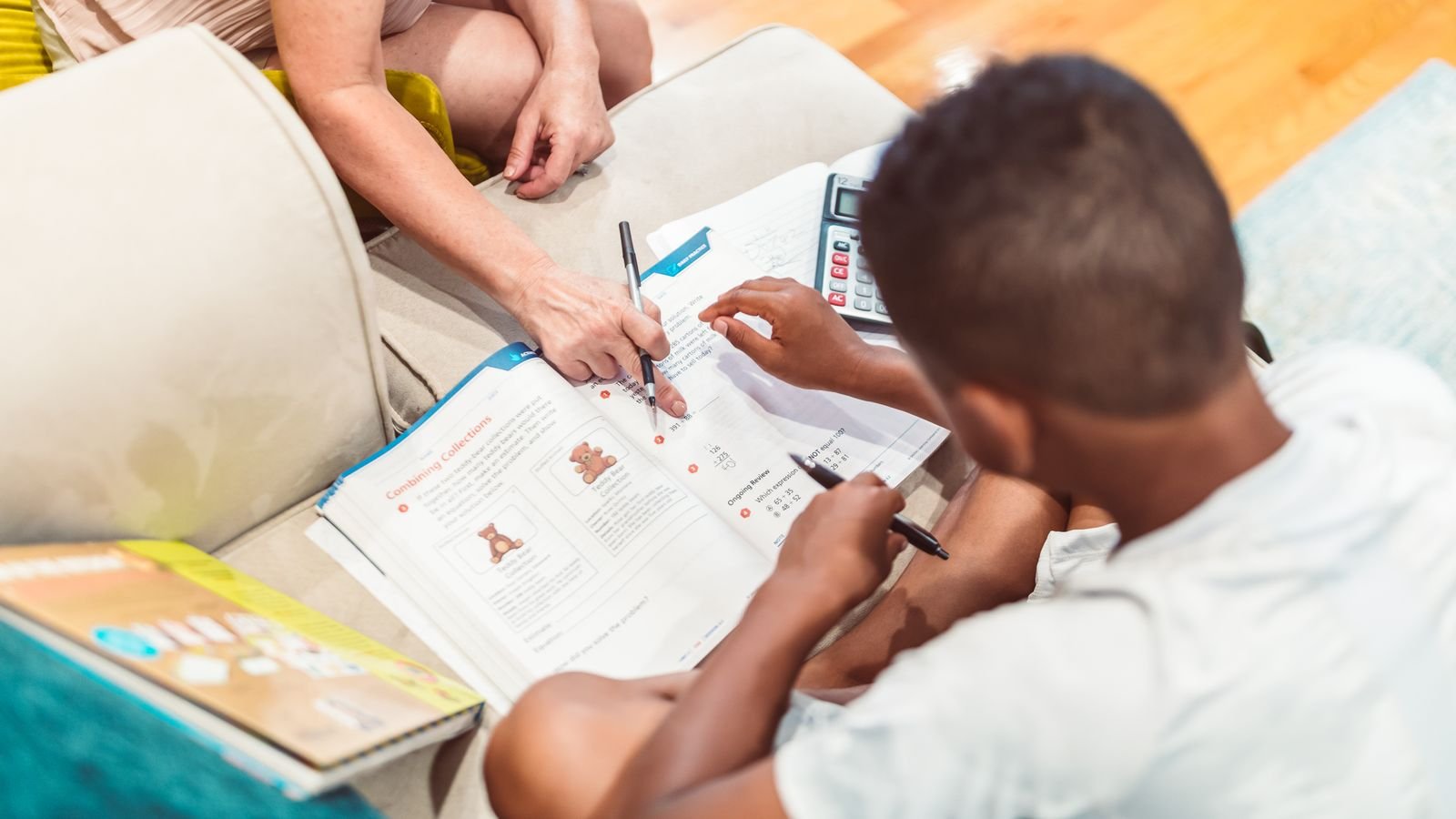 Parent supporting a primary school child working through a maths problem beside an open laptop during an online maths class