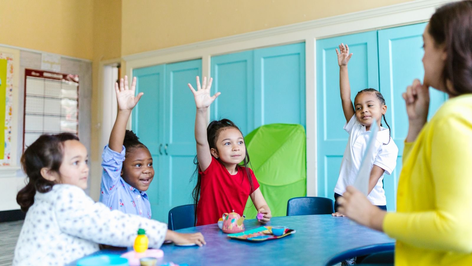 Group class of children raising hands while one quiet child waits for teacher attention